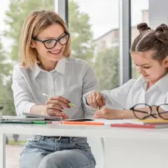 Kobieta z okularami uczy dziecko przy biurku, oboje w białych koszulach.