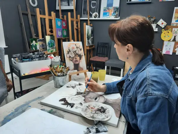 Kobieta w dżinsowej kurtce maluje obraz z kwiatami na płótnie w pracowni artystycznej.